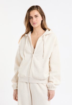 Kremowa bluza z kapturem wykonana z miękkiego materiału, wyposażona w zamek błyskawiczny, regulowany kaptur ze sznurkiem oraz ściągacze na rękawach, o luźnym kroju i gładkiej fakturze.