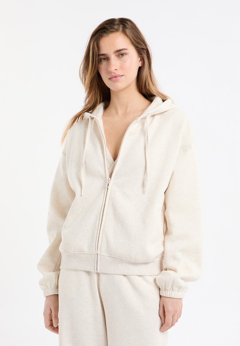 Sweat à capuche crème en tissu doux, avec fermeture éclair, capuche à cordon réglable et poignets côtelés, coupe décontractée et texture unie.
