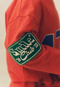 Czerwony bawełniany sweatshirt z dużą niebieską literą 'A' oraz zieloną haftowaną naszywką z napisem "Smile Club '19" na lewym rękawie.