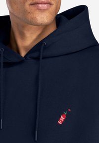 Sweat à capuche bleu marine en mélange de coton, avec un petit motif brodé de bouteille rouge et verte sur le côté gauche de la poitrine.