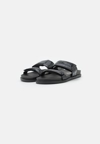 Svarta slidesandaler med texturerade remmar, platt sula och justerbara kardborrefästen; tillverkade av syntetmaterial för hållbarhet.
