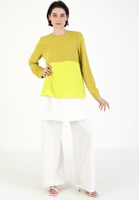Top a maniche lunghe in verde oliva e giallo brillante, abbinato a pantaloni bianchi larghi. Il materiale sembra morbido con una texture liscia.