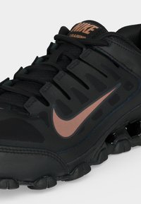 Μαύρο παπούτσι προπόνησης Nike με λογότυπο και κορδόνια σε ροζ χρυσό, με τμηματική σόλα σχεδιασμένη για ευκινησία και στήριξη.