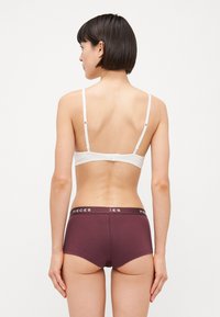 Bijela bralettica sa tankim trakama i podesivim kopčama; uparen s burgundskim boyshorts hlačicama koje imaju elastični pojas s "PIECES" logotipom.