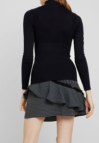Haut noir à manches longues côtelé avec un col montant, associé à une jupe courte à carreaux avec des détails à volants et une bordure en dentelle le long de l'ourlet.