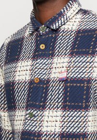 Camicia di flanella a quadri navy e crema con bottoni verdi e marroni, decorata con cuciture decorative e un piccolo etichetta sul lato sinistro del petto.