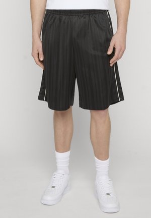 Person trägt schwarze gestreifte Sportshorts mit weißen Seitenstreifen, weiße Sportsocken und weiße Turnschuhe und steht vor einem schlichten Hintergrund.