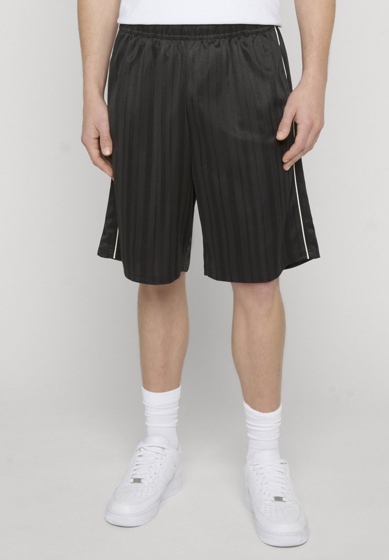 Personne portant un short de sport noir à rayures blanches sur les côtés, des chaussettes blanches montantes et des baskets blanches, debout devant un fond uni.