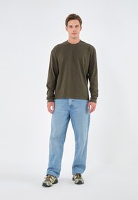 Olivengrünes Langarmhemd, hellblaue, locker sitzende Jeans und braune Camouflage-Schuhe, stehend vor einem weißen Hintergrund.