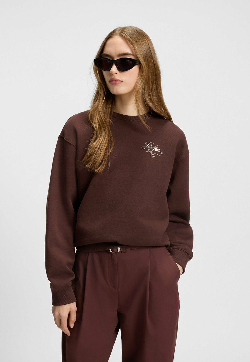 Felpa marrone con logo ricamato, vestibilità over-size, polsini a coste, abbinata a pantaloni scuri. Il modello indossa occhiali da sole neri a forma angolare.