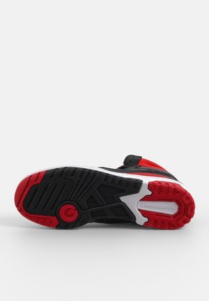 Semelle de chaussure de sport noire et rouge avec un motif de bande de roulement profond, semelle intermédiaire blanche et logo "new balance" sur un fond blanc propre.