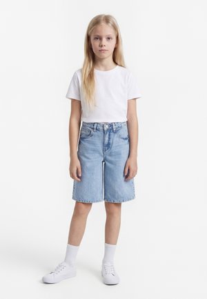 Niña joven con cabello largo y rubio, vestida con una camiseta blanca, pantalones cortos de mezclilla hasta la rodilla, calcetines blancos y zapatillas blancas, de pie contra un fondo liso.