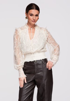 Vrouw draagt een doorschijnende witte kanten blouse met lange mouwen en zwarte leren broek met hoge taille, poserend tegen een lichte achtergrond.