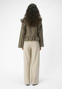 Olivgrüne Bluse mit Rüschendetails, langen Ärmeln und peplumförmigem Saum. Kombiniert mit cremefarbenen weit geschnittenen Hosen und schwarzen Schuhen.
