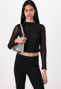 Črna prosojna majica z dolgimi rokavi in skrajšano obliko, v kombinaciji z visokimi črnimi hlačami. Model drži majhno kovinsko torbico.