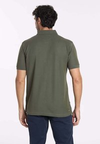 Polo shirt vert olive à manches courtes, texture lisse et col classique. Présente un petit logo sur l'ourlet inférieur.