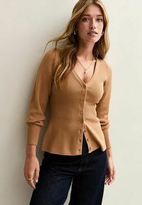 Mujer con cabello largo y rubio, vestida con un cárdigan beige de botones y pantalones oscuros, de pie contra una pared clara y lisa.