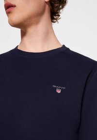 Mörkblå långärmad crewneck sweatshirt i bomull, med en broderad "GANT"-logotyp och sköld på bröstet. Slät textur.