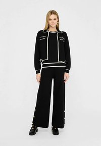 Czarny dziany top z białymi detalami wykończenia oraz pasujące szerokie spodnie; spodnie mają ozdobne guziki na nogawkach; do tego czarne buty.