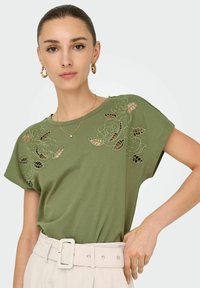 T-shirt vert olive avec design découpé floral sur les manches. Tissu doux, coupe décontractée, associé à une ceinture crème. Forme simple et moderne.