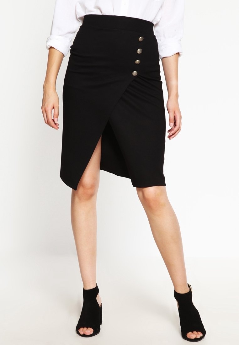 Femme portant une jupe asymétrique noire avec des boutons latéraux et une fente à l'avant, associée à des bottines noires à talons ouvertes et une chemise blanche.