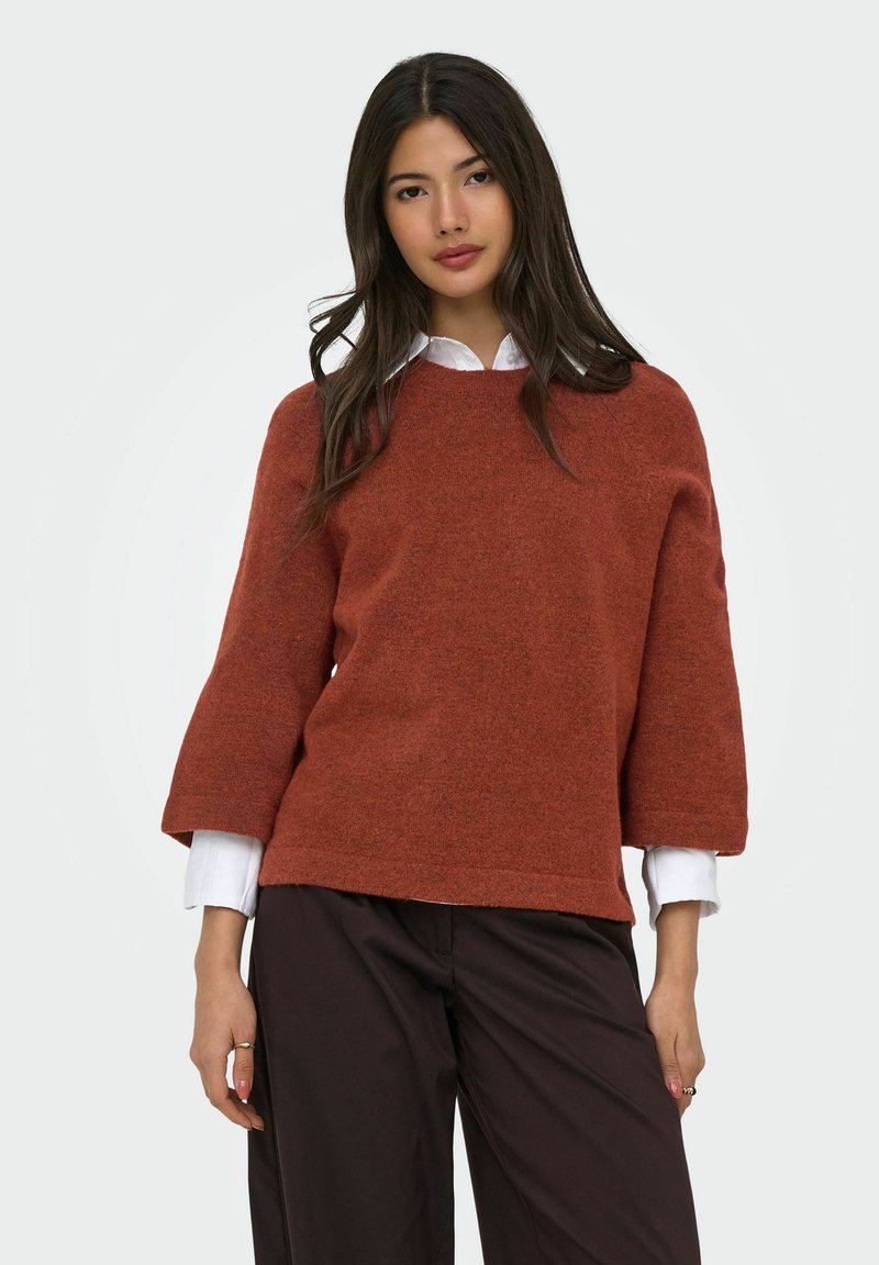 Femme aux longs cheveux foncés portant un pull couleur rouille par-dessus une chemise blanche et un pantalon marron foncé, debout devant un fond clair uni.