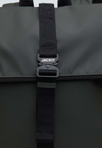 Tumma harmaa reppu, jossa on sileä, teksturoitu pinta. Varustettu mustalla hihnalla, jossa on kohokuvioitu teksti ja kestävä mustasta muovista valmistettu solki.