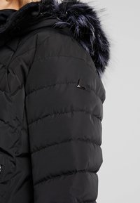 Manteau d'hiver noir matelassé avec une garniture en fourrure autour de la capuche et un petit logo métallique sur le haut du bras, porté par une personne.