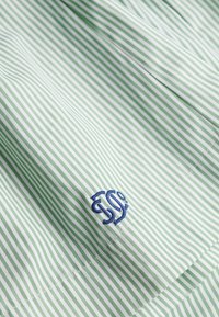 Tissu rayé vert et blanc avec une texture lisse, présentant un logo brodé bleu sur le côté inférieur droit.