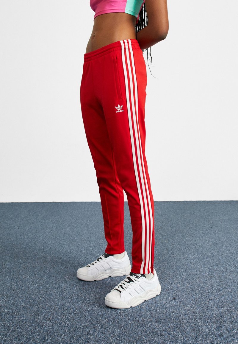 Rote Adidas Jogginghose mit weißen Seitenstreifen, elastischem Bund und Seitentaschen, kombiniert mit weißen Turnschuhen auf einem strukturierten blauen Boden.