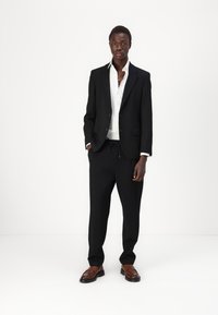 Veste de costume noire ajustée sur une chemise blanche, associée à un pantalon noir avec une ceinture élastique, et des chaussures en cuir marron.