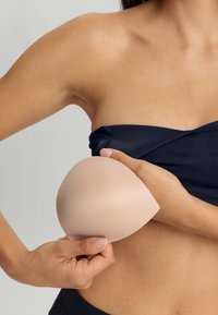 Beige silikone bh-kop holdt i hånden, glat tekstur, oval form, parret med en marineblå top, der viser hudtone baggrund.