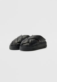 Schwarze Leder-Slipper mit geflochtenem Design auf der Vorderseite und einer dicken, strukturierten Sohle. Minimale Markenkennzeichnung an der Seite.