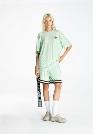 Persona rubia con camiseta verde claro texturizada de gran tamaño y pantalones cortos a juego con ribete negro, calcetines blancos y zapatos beige texturizados sin cordones.