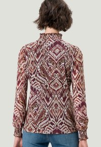 Chemisier à manches longues en tissu à motifs avec des teintes violettes, blanches et vertes ; col et poignets froncés ; coupe décontractée ; design géométrique complexe.