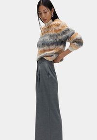 Flauschiger gestreifter Pullover in Orange, Grau und Creme, kombiniert mit hoch taillierten grauen Hosen mit Falten, die einen Lagenlook erzeugen.