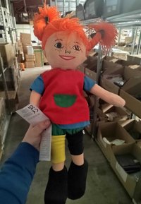 Doll with orange hair styled in pigtails, wearing a red vest, blue shirt, green shorts, and black boots. Tag visible in hand.