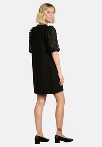 Czarna sukienka z przezroczystymi, bufiastymi koronkowymi rękawami. Krótkie, proste cięcie do kolan. Modelka ma na sobie czarne szpilki. Prosta i elegancka sylwetka.