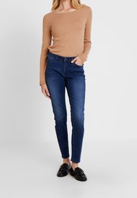 Mujer vistiendo un suéter largo de manga larga color camello, jeans ajustados oscuros y mocasines negros con flecos, de pie con una mano en el bolsillo.
