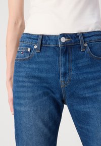Ingrandimento di una persona che indossa jeans di denim blu e una maglietta bianca, evidenziando le tasche frontali, il bottone e i dettagli delle cuciture.