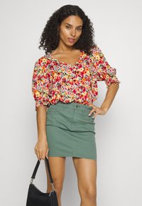 Blusa floral com mangas bufantes em rosa, vermelho e amarelo, combinada com uma saia de ganga verde. Modelo leva uma mala preta.