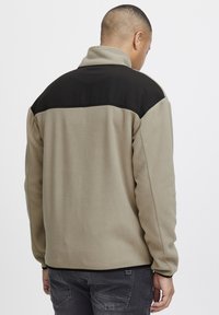 Chaqueta de forro polar en beige claro con un panel negro en los hombros. Presenta un cuello alto y una textura suave. Vista trasera, mostrando los detalles de costura.
