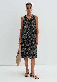 Schwarzes ärmelloses Kleid mit weißen Punkten, V-Ausschnitt, fließende A-Linie, getragen mit einer beigen Tragetasche und modischen Sandalen.
