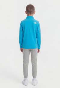 Αγόρι που στέκεται γυρισμένο προς τα πίσω, φοράει ένα φωτεινό μπλε φλις μπουφάν, ανοιχτό γκρι φόρμα, λευκές κάλτσες και λευκά αθλητικά παπούτσια σε λευκό φόντο.