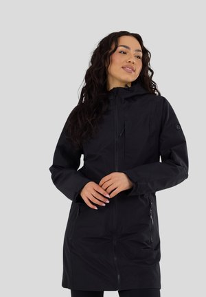 Donna con lunghi capelli ricci che indossa un cappotto nero con cappuccio e cerniera, posa con le mani intrecciate davanti a uno sfondo neutro.