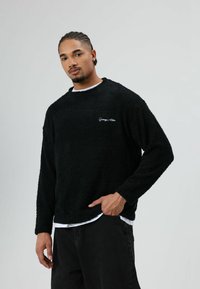 Zwarte pluche trui met lange mouwen en ronde hals, met een wit geborduurd logo op de borst, gedragen over een wit overhemd.