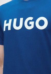 Μπλε μπλουζάκι με έντονη λευκή επιγραφή "HUGO" στο στήθος, φορεμένο από ένα άτομο.