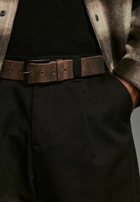 Ceinture en cuir brun avec une finition texturée, dotée d'une boucle en métal noir, portée sur un pantalon noir et un pull côtelé foncé.
