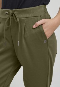 Olive krāsas jogger bikses, kas izgatavotas no mīksta auduma, ar auklu jostasvietā, sānu kabatām un nelielu metāla logotipu kabatas augšdaļā.