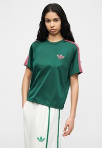 Νεαρή γυναίκα που φοράει πράσινο πουκάμισο Adidas με ροζ ρίγες και λογότυπο, σε συνδυασμό με λευκό παντελόνι Adidas με πράσινες ρίγες και λογότυπο.